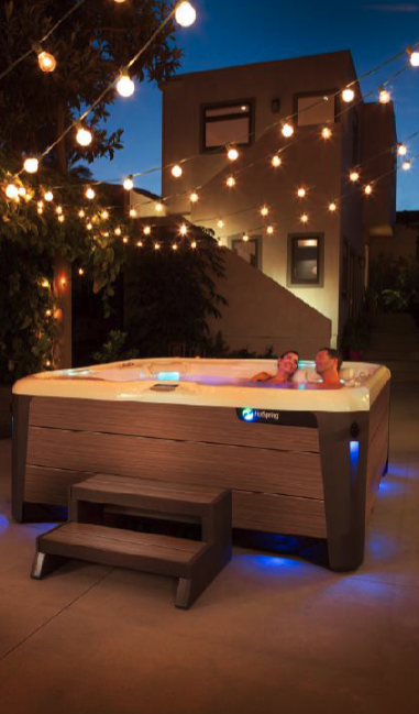Two people relax in a hot tub under string lights in a cozy, modern backyard setting during the evening.
